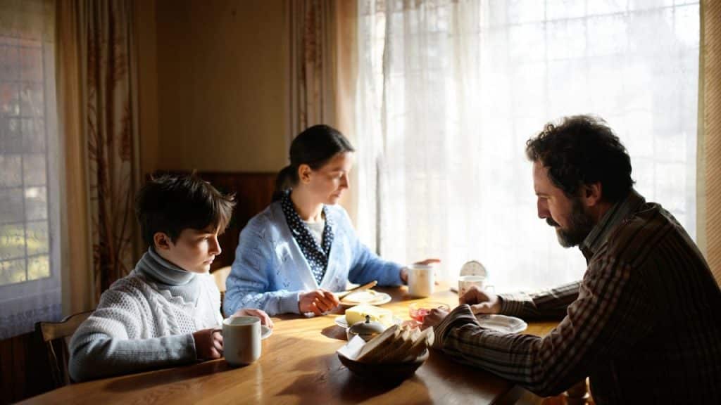 A family of three sitting at a dining table having a quiet meal in a sunlit room.