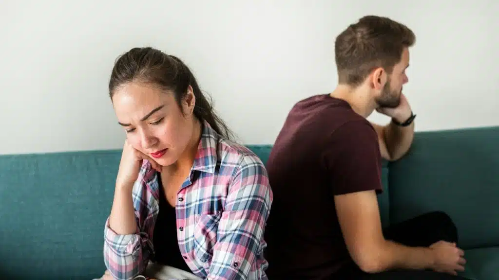 A woman with a worried expression sits facing forward on a couch with a pillow, while a man sits turned away from her in the background.