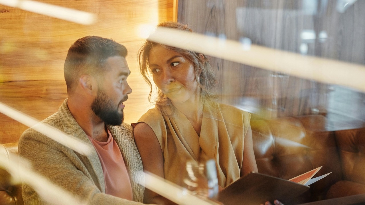 A man and woman sit together in a booth, viewed through a reflective glass surface.