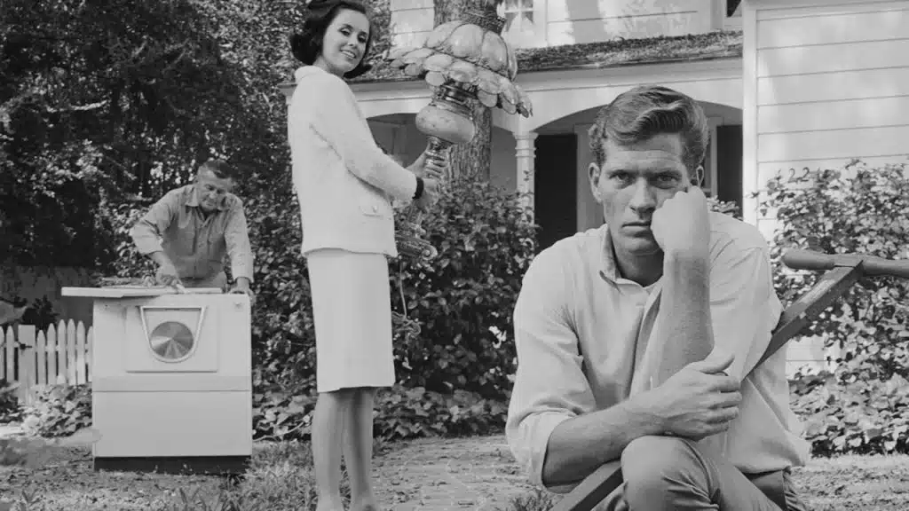 A bored man kneeling down next to a wheelbarrow while a woman standing behind is looking at him.