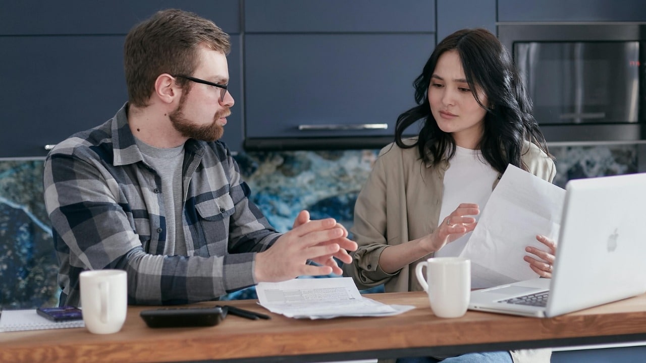 A man and woman sit at a counter with a laptop and papers, appearing to talk.