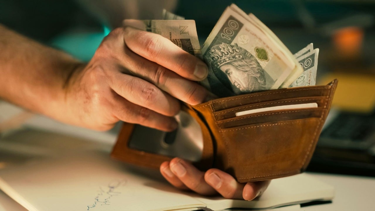A close-up of hands pulling paper bills out of a brown leather folding wallet.