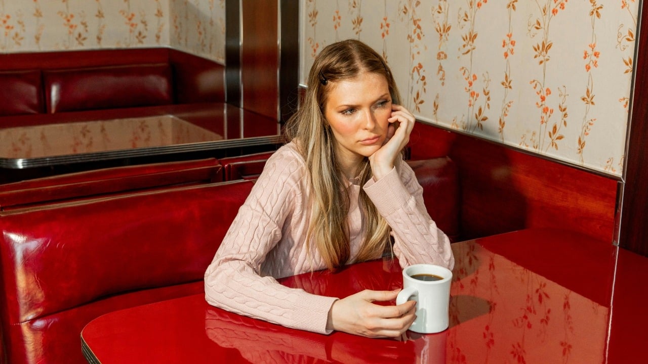 A woman in a pink sweater sits alone at a red booth holding a mug.