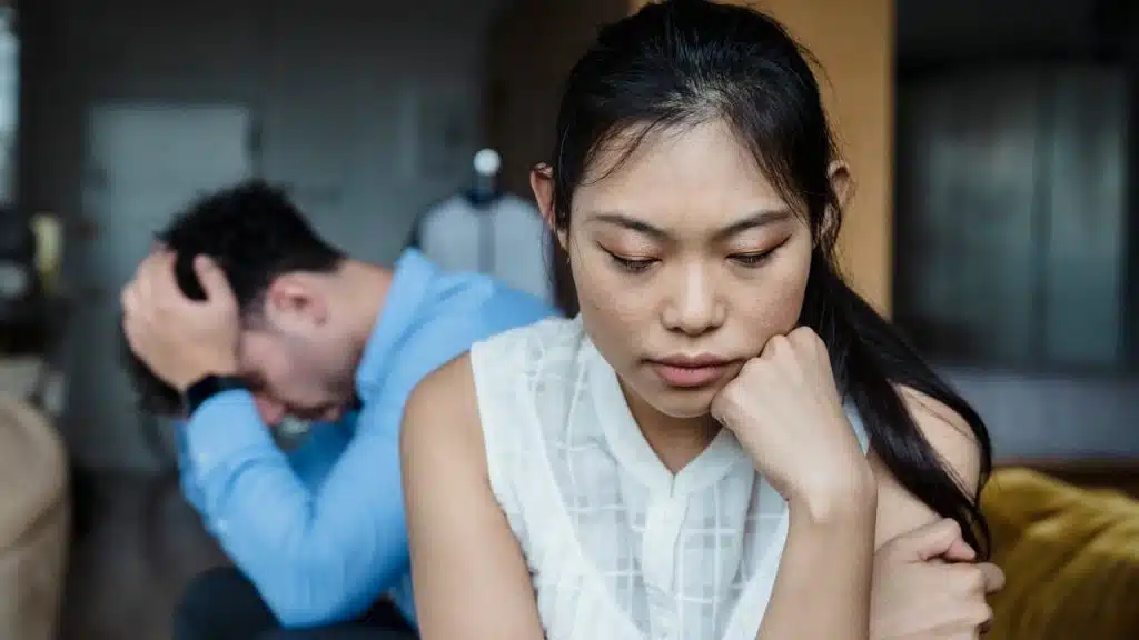 A distressed woman is sitting in the foreground with her hand on her chin, while a man covers his head in the blurry background.