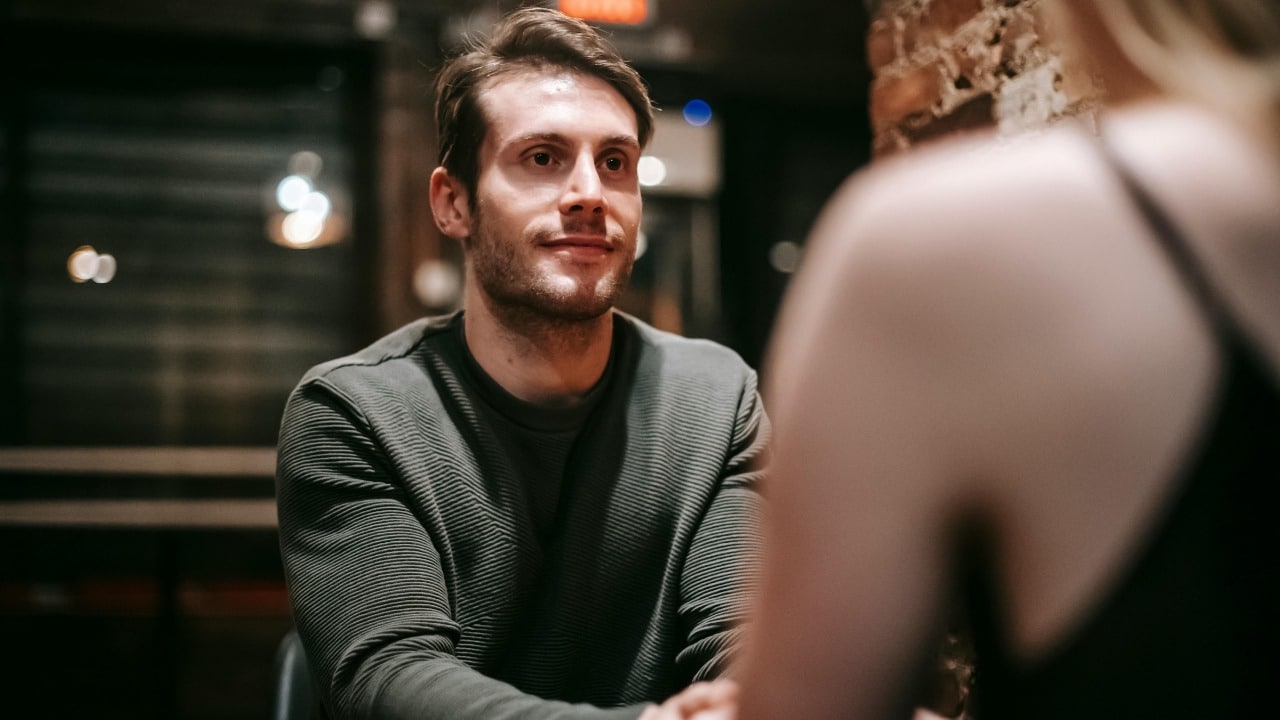 A man in a grey sweater looks forward while sitting across from a blurred person.
