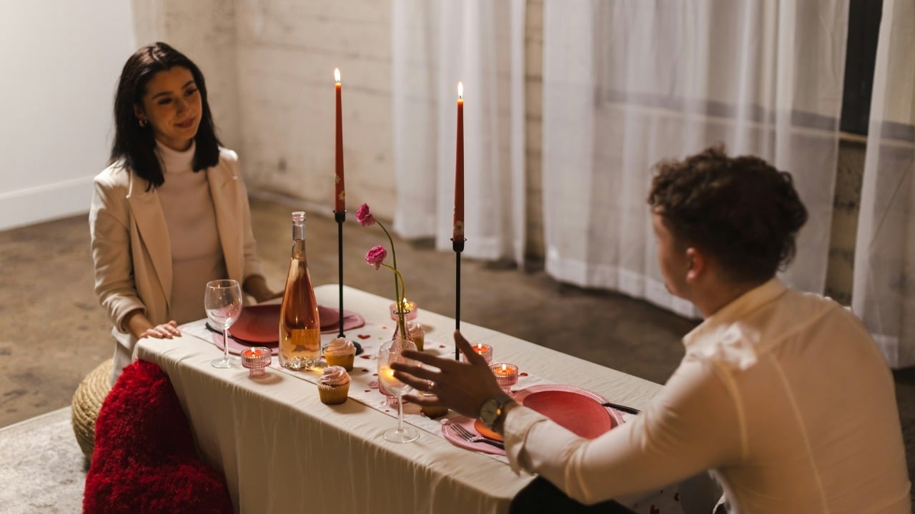 A man and woman sit at a candlelit table with wine, cupcakes, and red plates.
