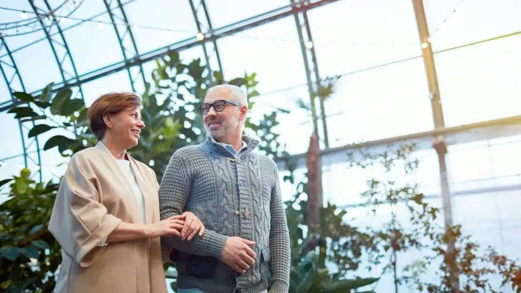 An older man in a gray sweater and a woman in a light jacket walking arm-in-arm through a sunny, tropical-like indoor garden.
