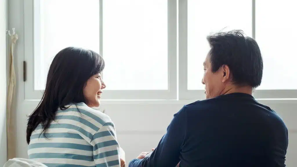 A man and woman sitting with their backs to a large, bright window, looking at and smiling at each other.
