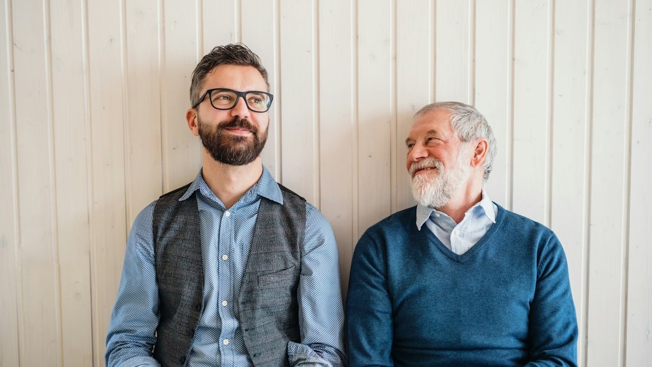 A bearded man wearing glasses sits next to an older man who stares at him.