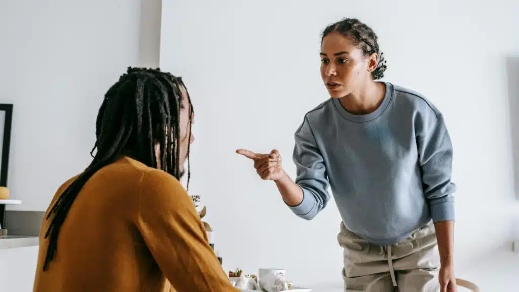 A woman stands and points a finger while speaking to a man sitting at a table.
