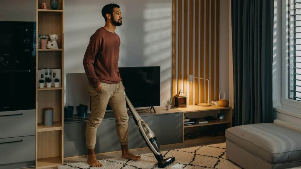 A man stands in a living room holding a vacuum cleaner while looking toward a window.