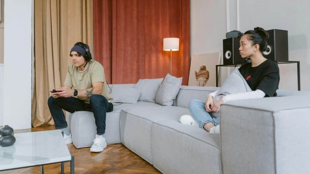 A man wears headphones and plays video games while a woman sits on the couch nearby.