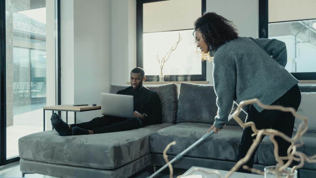 A man sits on a grey sofa with a laptop while a woman vacuums the floor.