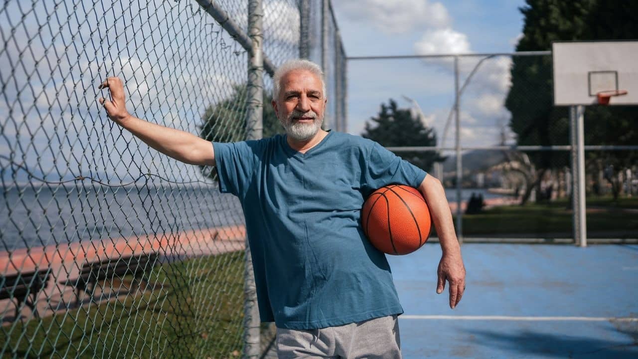 An older man with gray hair holds a basketball while standing next to a chain-link fence.
