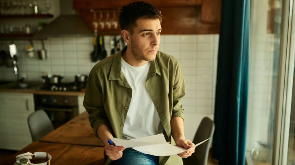 A man stands in a kitchen holding papers and a pen while looking out a window.