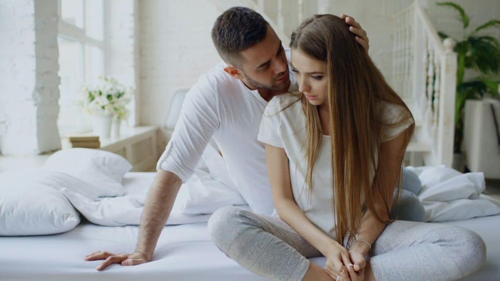 A man sits on a bed and places a hand on the head of a woman.