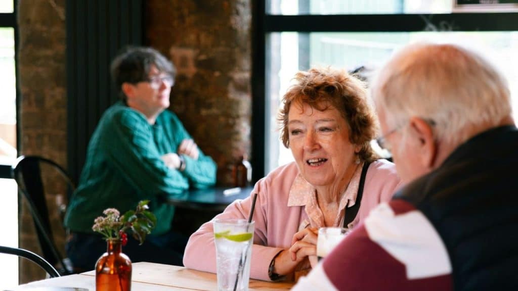 An older woman in a pink cardigan sits at a table talking to two people.