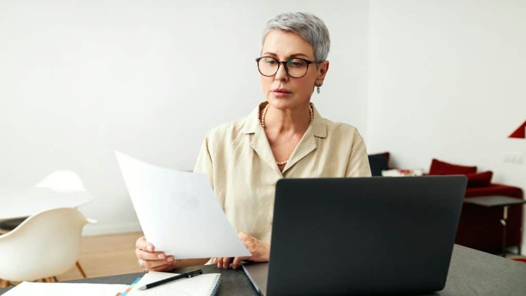 A woman with short grey hair and glasses looks at a paper held near a laptop.