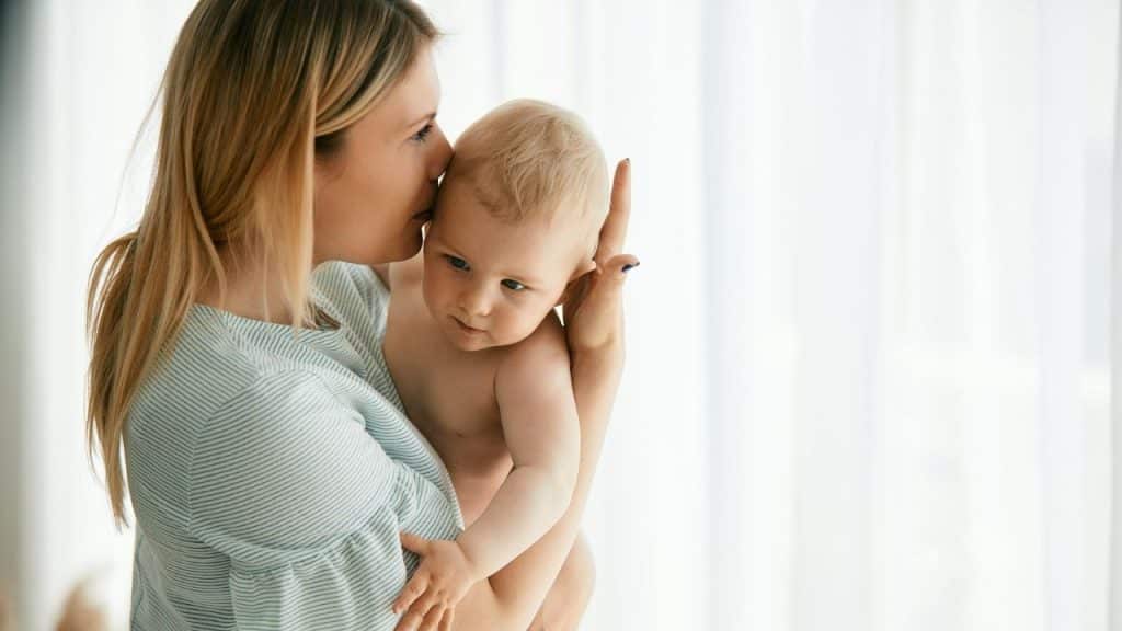 A blonde woman holds a baby close to her face in front of a white curtain.