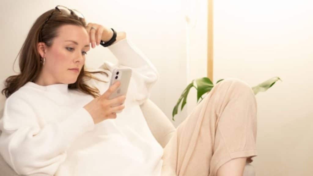 A woman in a white sweater leans back and looks at a white smartphone.
