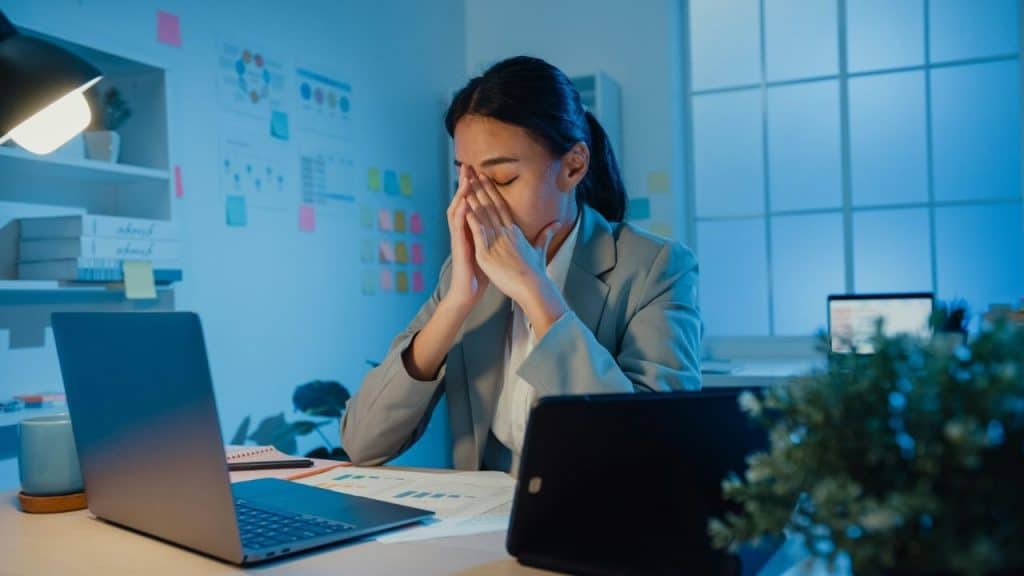 A woman in a grey blazer sits at a desk and covers her face with hands.