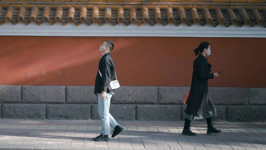 Two people walk in opposite directions on a sidewalk in front of a long red wall.