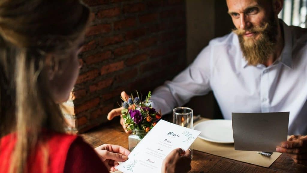 A man with a beard looks at a woman holding a menu at a wooden table.