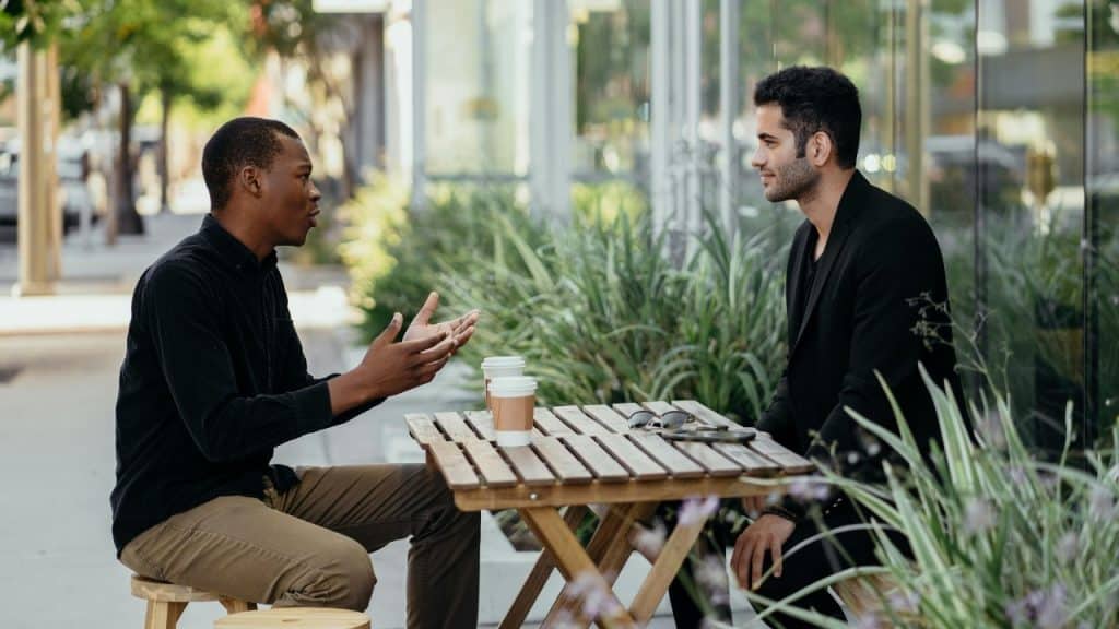 Two men sit at an outdoor wooden table with coffee cups, engaged in a serious conversation.