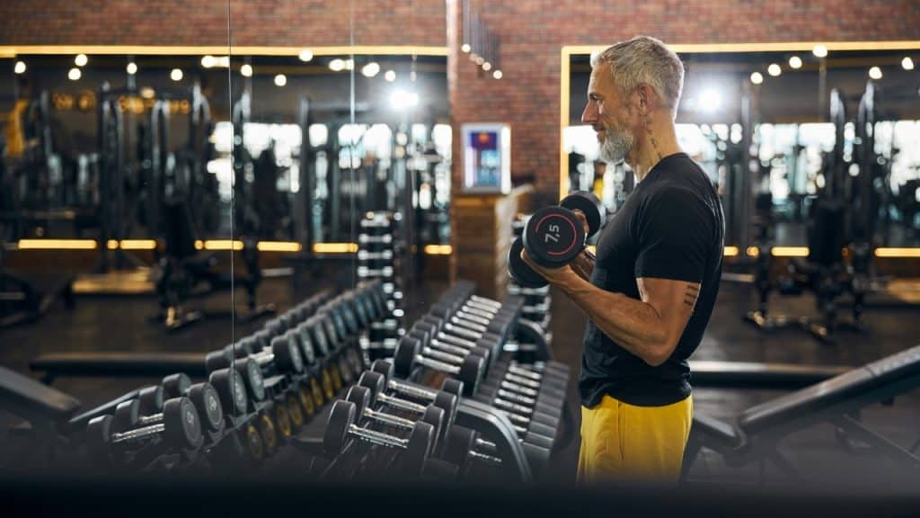 A fit man with grey hair performs bicep curls with dumbbells in a well-lit gym.