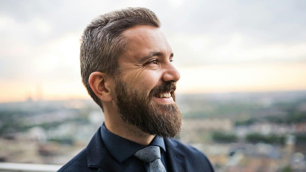 A close-up profile of a smiling man with a beard, wearing a suit and a tie.