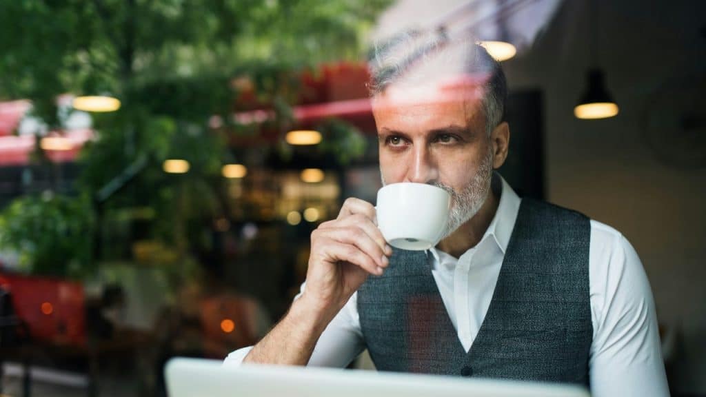 A man with grey hair and a vest drinks from a white cup behind a window reflection.