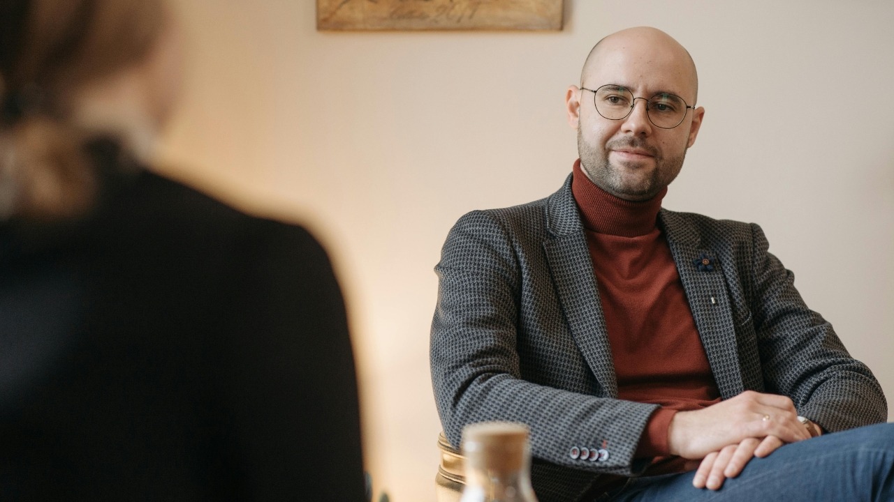 A bald man wearing glasses and a blazer sits in a chair during a focused conversation.