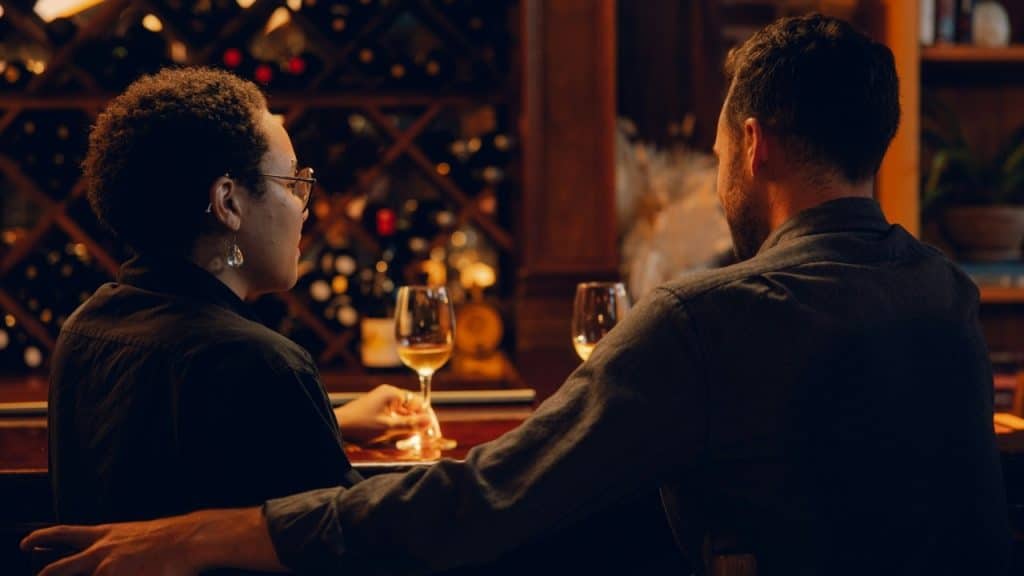 A man and woman sit at a dark wooden bar with wine glasses in front of them.