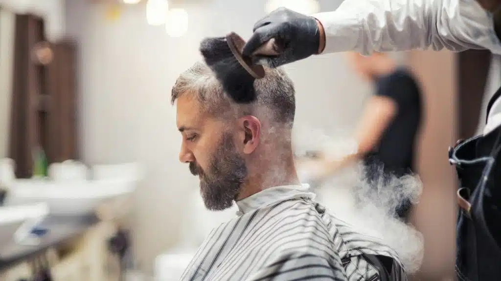 A barber uses a brush and powder on the neck of a man with a beard.