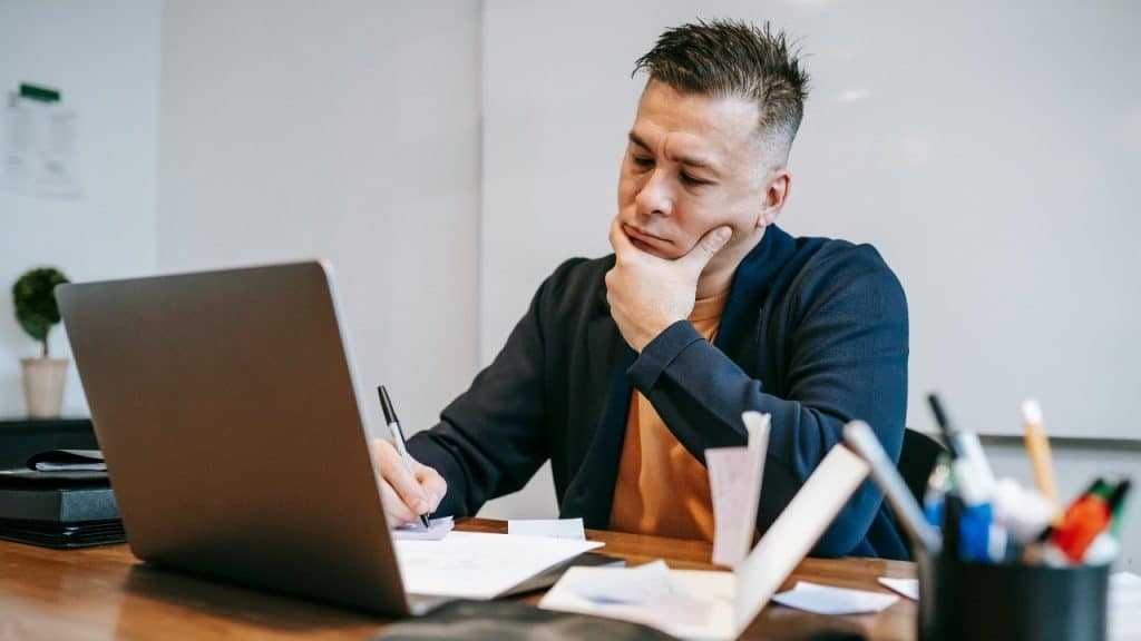 A man sits at a desk with a laptop while resting his chin on his hand.