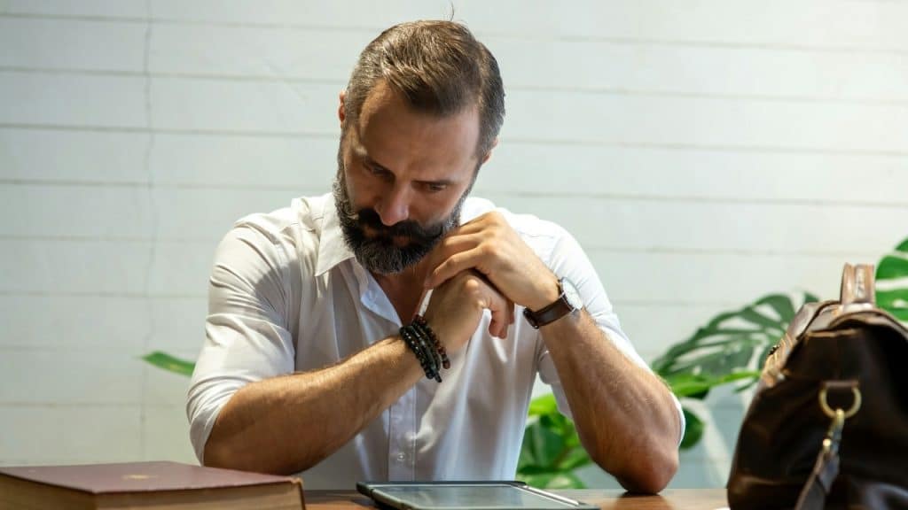 A man with a beard sits at a table with his hands clasped near a tablet.