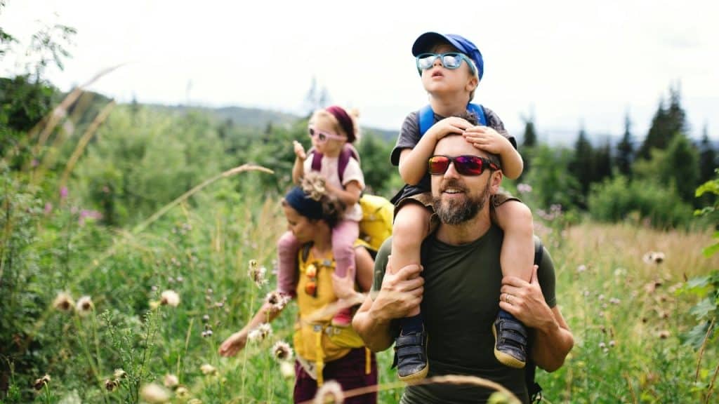 A man carries a child on his shoulders while walking through a tall grassy field.