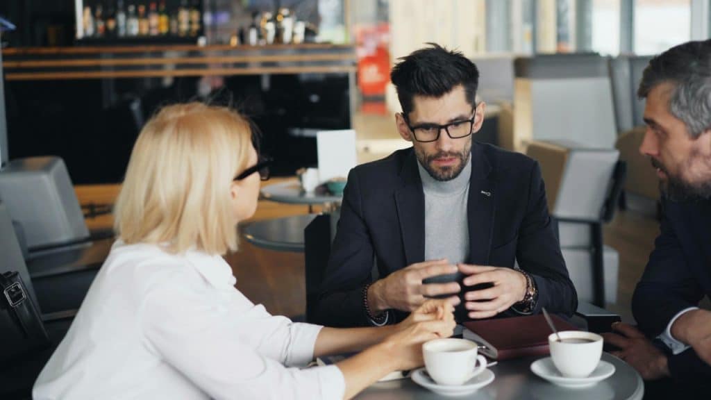 Three people sit at a table with coffee cups while a man gestures with his hands.