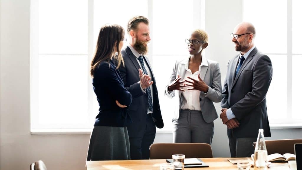 Four professionals in business attire stand in an office and engage in a group conversation.