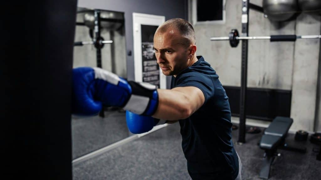 A man wearing blue boxing gloves punches a black heavy bag in a training gym.