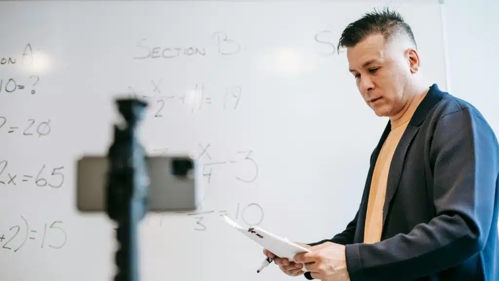 A teacher stands in front of a whiteboard holding papers.