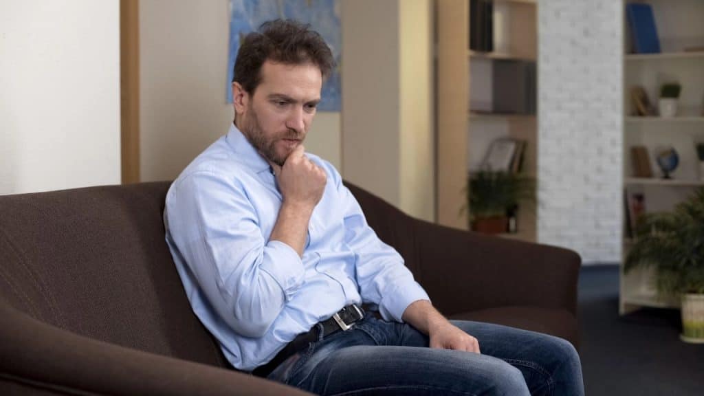 A bearded man in a blue shirt sits on a brown sofa, resting his chin on his fist.