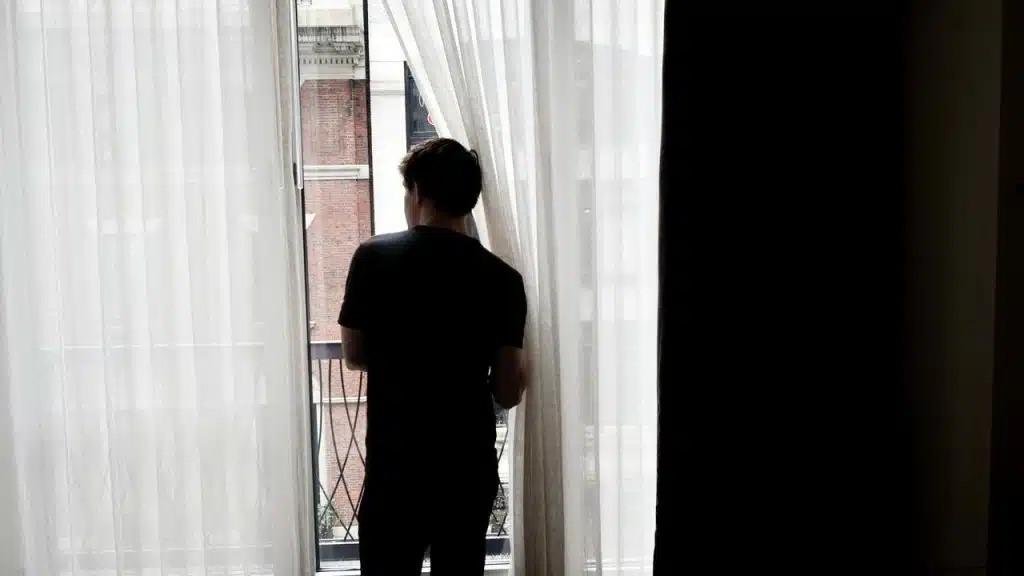 A person standing by a window, looking outside through sheer curtains.