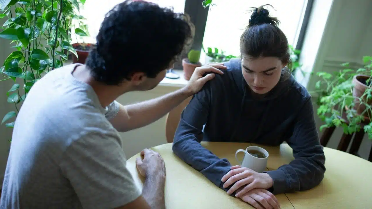 A man with a grey shirt comforts a woman in a black hoodie at a table.
