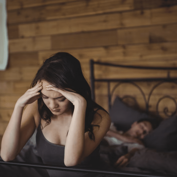 Upset woman sitting on a bed while man sleeping in background