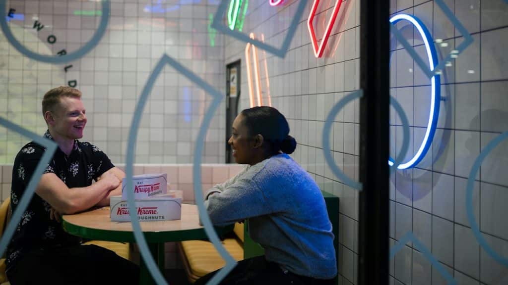 A smiling man and woman are sitting across from each other at a table in a modern restaurant or donut shop, with neon lights in the background.