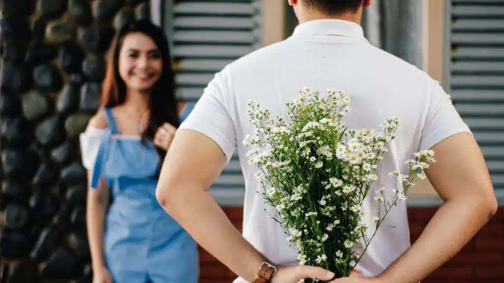 A man stands with his back to the camera, holding a bouquet of white flowers behind him, while a smiling woman in a blue outfit stands blurred in the background.