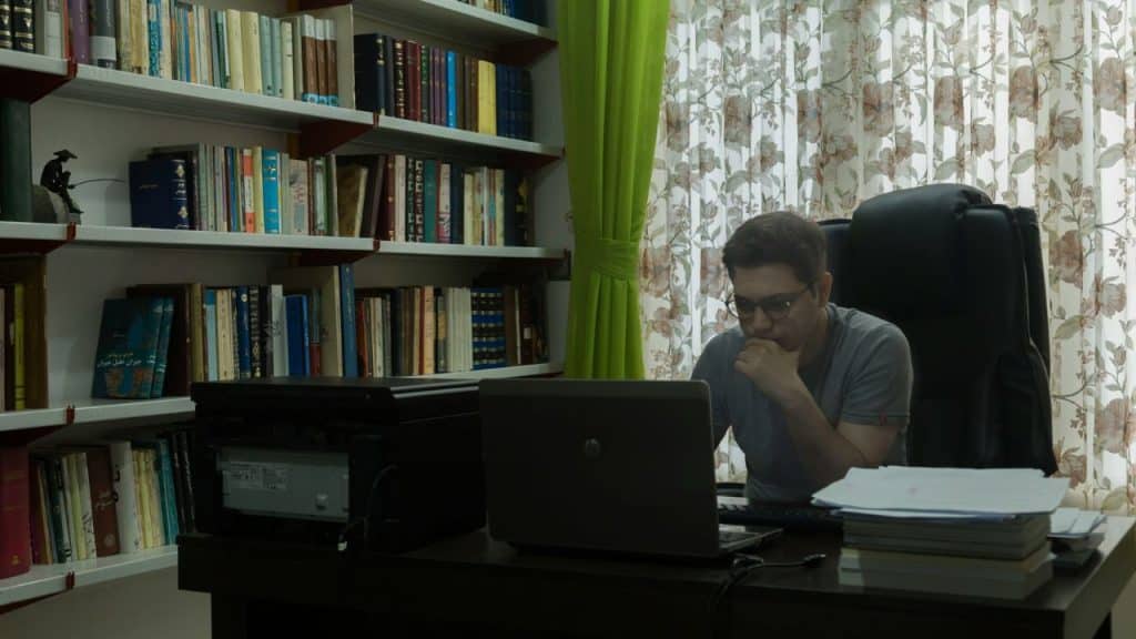 A man with glasses sits at a desk, looking intently at his laptop.
