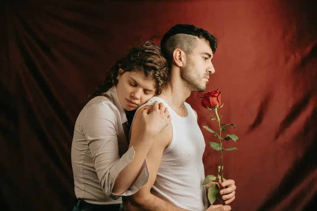 A woman leaning on the shoulders of a man while he holds a rose with a sad face.