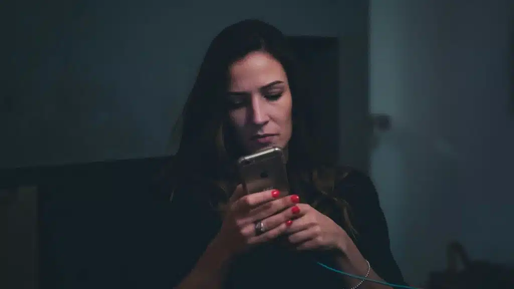 A woman with red nail polish looks intently at her phone in a dimly lit room.